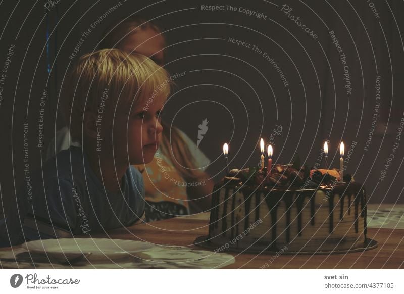 A blond boy is sitting at a table in a dark room and looking at a birthday cake with burning candles. Siblings are waiting for dessert at the birthday celebration.