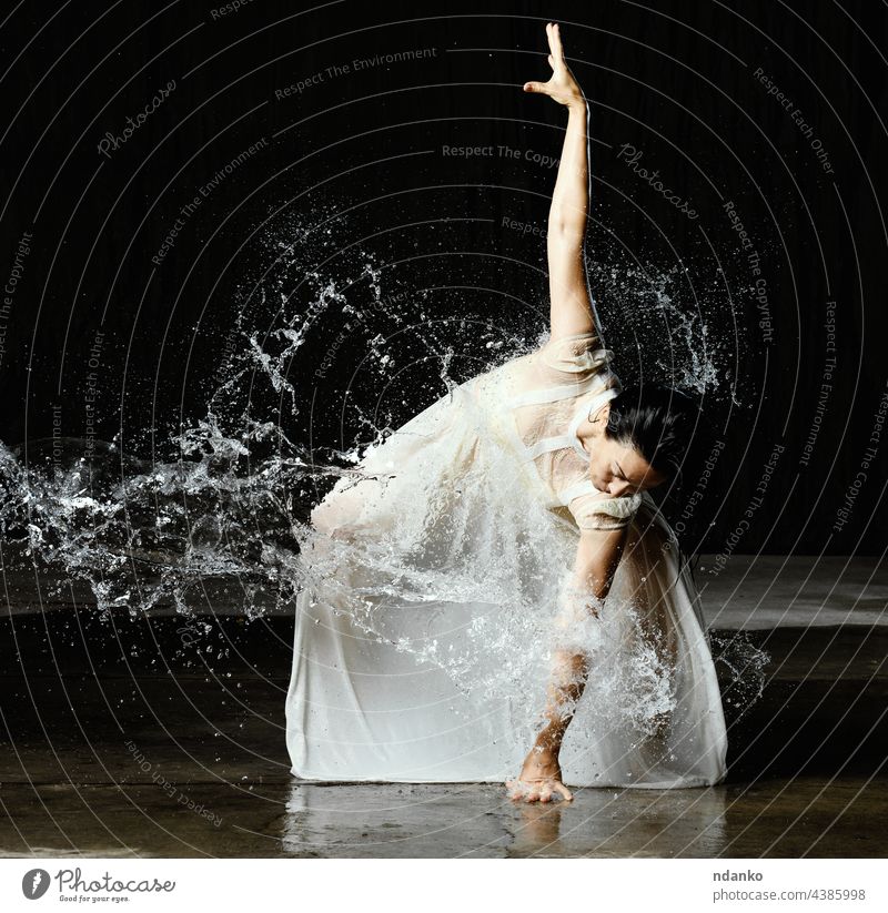 beautiful woman of Caucasian appearance with black hair dances in drops of water on a black background. The woman is wearing a white chiffon dress flexible aqua