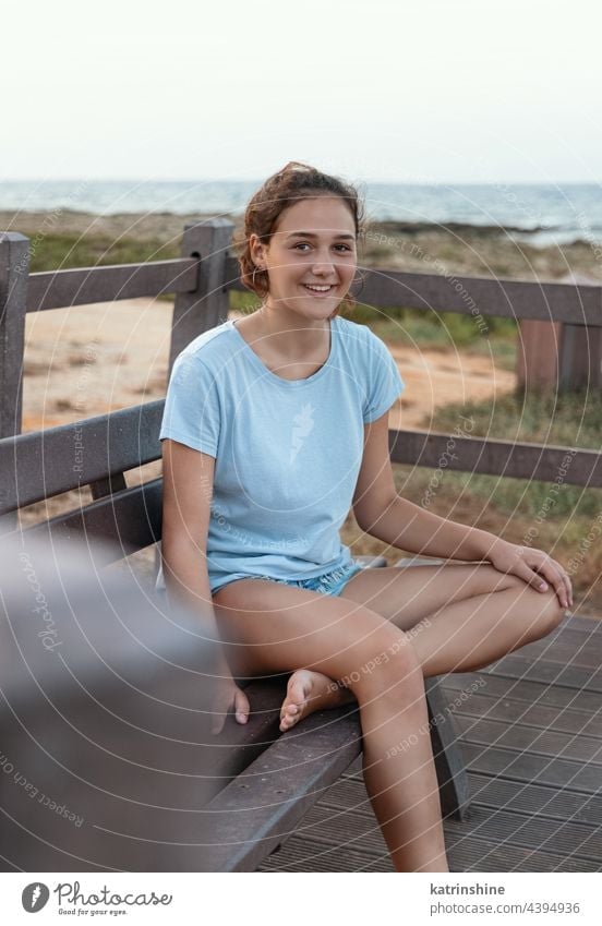 Smiling teenage girl sitting on the wooden bench near the coast outdoor teenager sea sunset mockup blue Caucasian adolescent cliff teen girl wearing t-shirts
