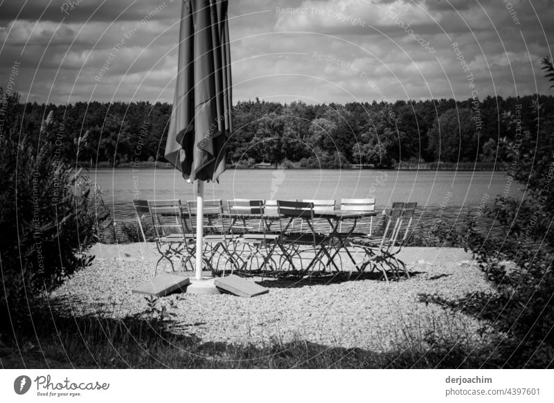 A folded parasol, a few empty chairs and table, and the water from a lake and in the background a lot of forest. To everything in the sky many clouds. Now only the guests are missing...