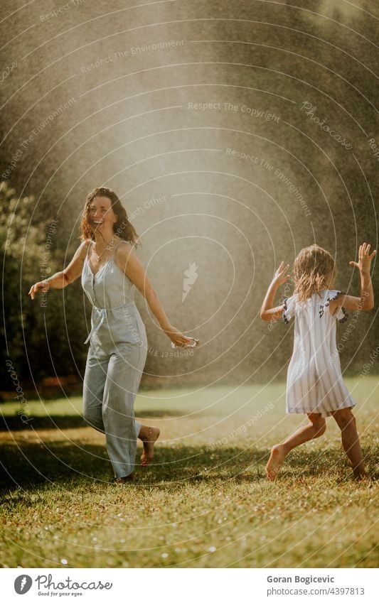 Cute little girl having fun under irrigation sprinkler with her mother active activity amusement backyard cheerful child childhood day drops enjoy freedom funny