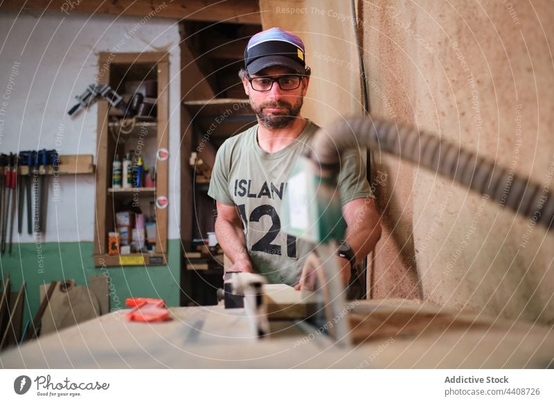 Focused carpenter cutting lumber on sawing machine in workroom plank precise workshop man craftsman woodwork small business concentrate accuracy eyewear hose