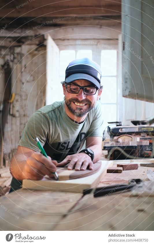Smiling craftsman with marker and wooden blocks on workbench trace skateboard manufacture plank woodwork accuracy cheerful handmade small business joiner