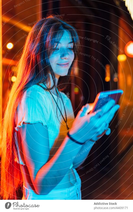 Smiling woman chatting on smartphone in neon lights interested internet online spare time town using gadget device watching cellphone gentle sincere charming