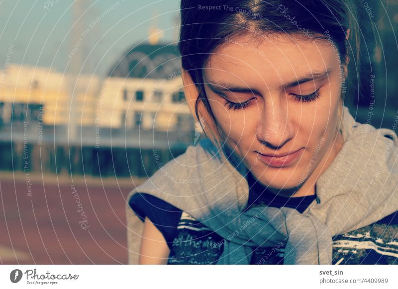 Urban portrait of a girl with her eyes lowered because of the bright sun. A nice girl outdoors against the background of a fountain in a summer evening.