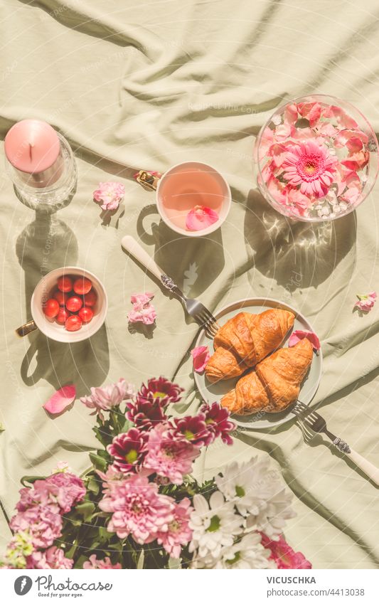 Sunny aesthetic picnic breakfast with flowers floating in crystal vases, croissants with tea from rose petals , bouquet of flowers and candles on blanket. Top view. Outdoor