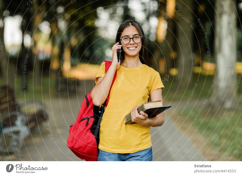 Portrait of a beautiful brunette with glasses and a backpack talking on the phone in the park girl student walking campus pretty books smiling holding young