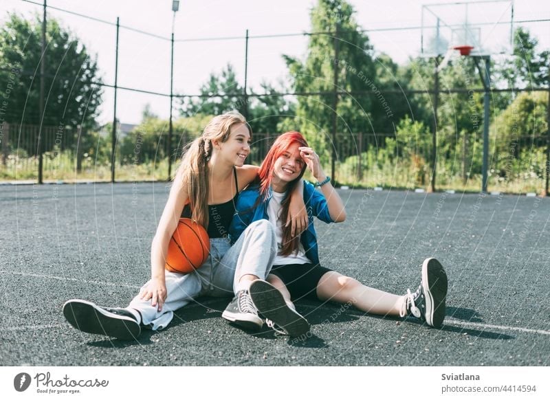 Two girls in sports clothes and with a basketball are chatting, sitting on the playground. Sports, competition, friendship teenager court team best friends