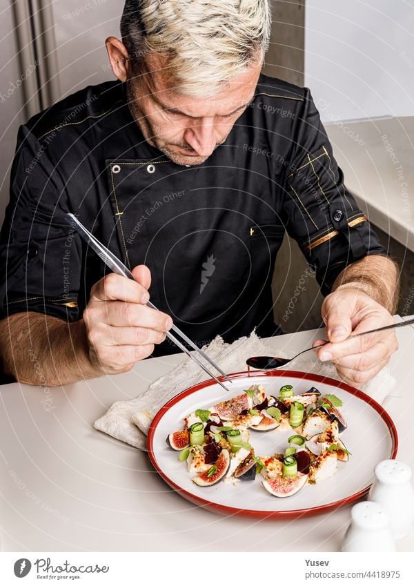 A stylish chef in a black jacket prepares tasty salad with ripe figs, goat Chevre cheese, fresh cucumber roll, beetroot and mustard leaves with tomato sauce. Restaurant dish. Delicious food