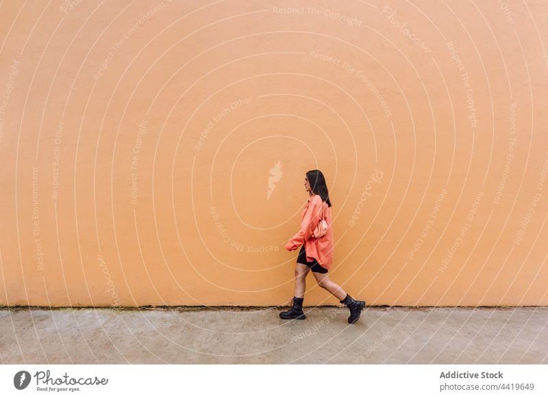 Young woman walking near orange wall cool style modern street trendy millennial individuality confident appearance female sunglasses stroll brunette long hair