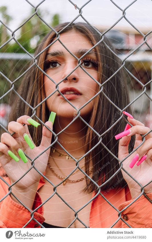 Young woman standing behind metal grid millennial portrait trendy cool style manicure long confident female charming feminine interest gaze serious brunette