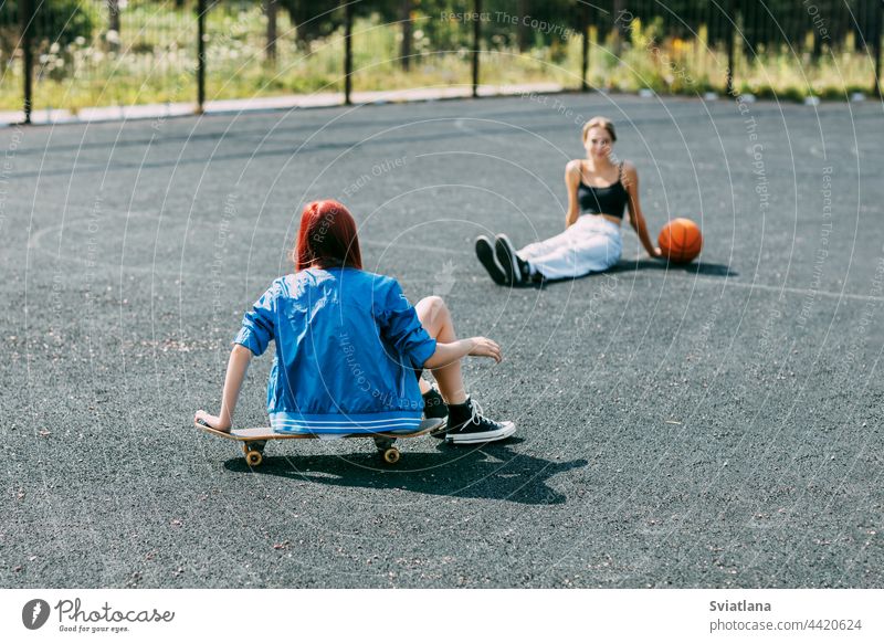 A young girl is sitting on a skateboard outdoors on a basketball court with her basketball player friend skateboarder sport skateboarding skating teenage ride