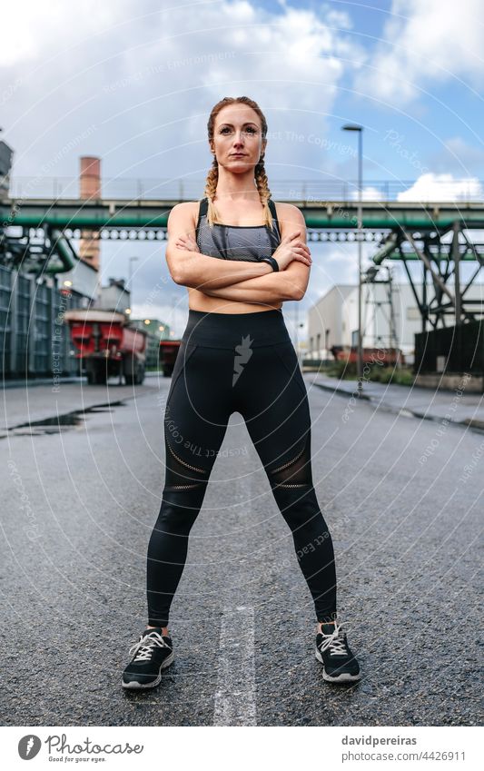 Sportswoman with crossed arms posing in front of a factory sportswoman strong self-confident looking straight ahead runner clouds serious empowered