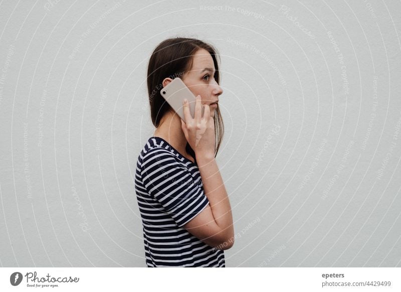 Young woman holding a smartphone to the ear and standing in front of a white wall brown hair casual clothes copy space gen z getting news millenial