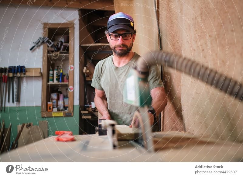 Focused carpenter cutting lumber on sawing machine in workroom plank precise workshop man craftsman woodwork small business concentrate accuracy eyewear hose