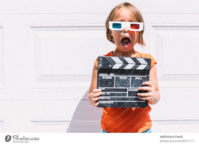 Trendy kid in 3D glasses holding clapperboard standing on wall three dimensional amazed entertain 3d fun eyewear joy astonish child casual girl watch actress