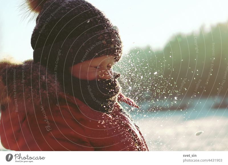 Snowflakes sparkle in the sunlight in the air in front of the face of a child who has closed his eyes. A boy in winter clothes plays with snow outdoors on a frosty sunny winter day.