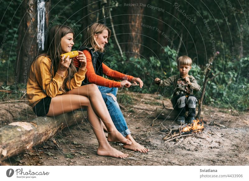 A young mother with her children is sitting by the campfire during a joint holiday, a girl is eating corn camping mom kids weekend hike daughter son together