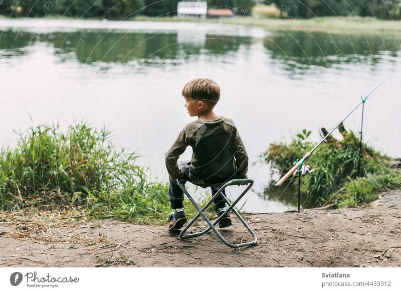 The boy is sitting on a folding chair on the shore of a lake or