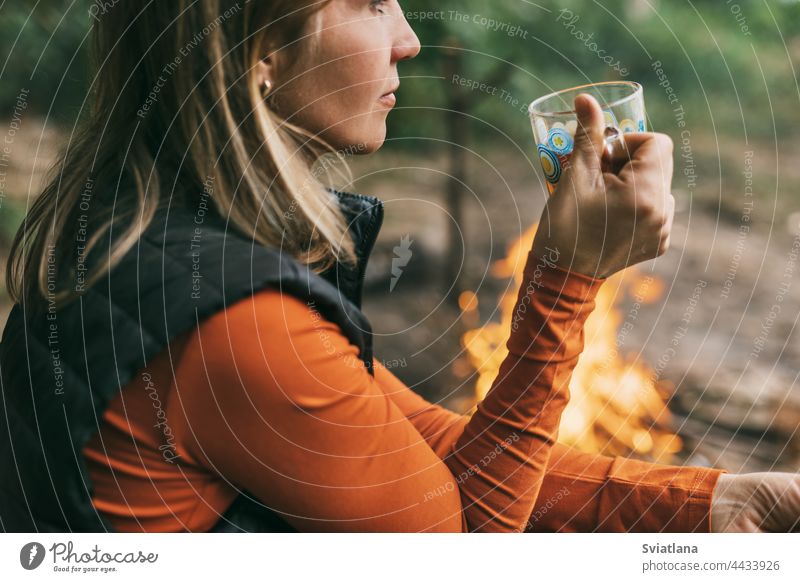 Close-up of an adult woman sitting with a cup of tea by the fire while relaxing in the forest camping drink young nature tourist adventure vacation campfire