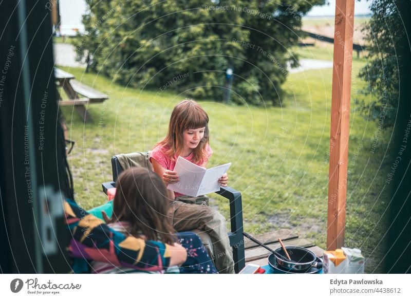 Girl reading in front of the wooden cabin at the campsite real people authentic mother parent together love house children family kids camping field rural green