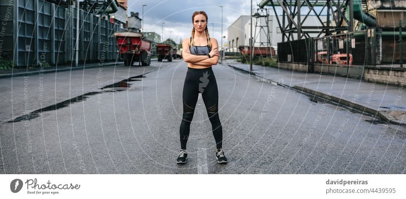Sportswoman looking camera posing in front of a factory sportswoman runner middle road asphalt self-confident crossed arms banner web header panorama panoramic