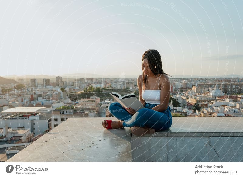 Cool black woman reading book on rooftop in city textbook spare time literature trendy legs crossed attentive sandal creative design footwear focus millennial