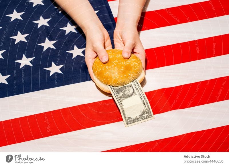 Crop person with cut bun and banknote above USA flag dollar price hamburger money fast food concept american cost 4th july independence day usa national