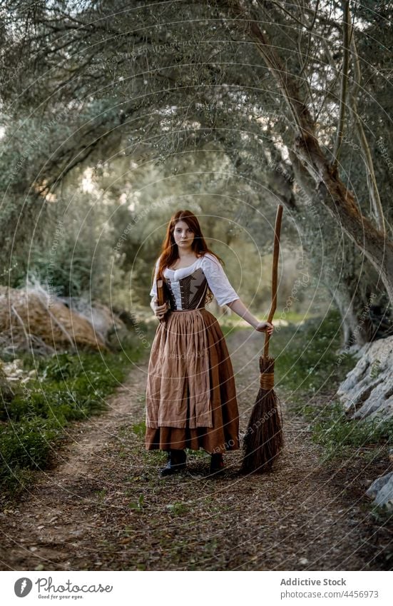 Witch with broomstick and book in forest witch magic woman dress enchantress autumn mystic female witchcraft magician woods stand dull gloomy halloween serious