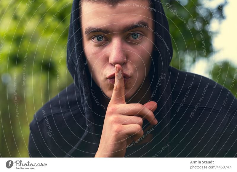 be quiet, portrait of a young man holding a finger to his mouth gesture expression hand eyes hoodie closeup evening bokeh nature silence secret silent mute