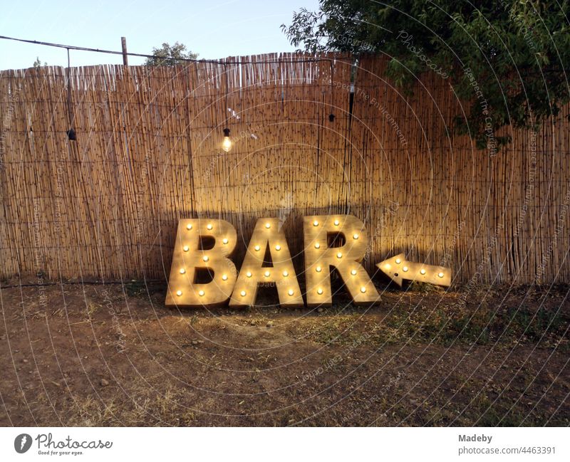 Illuminated indication of a bar in natural colors in front of color-matching brushwood fence in summer at dusk at a Turkish wedding in the park in Güzelbahce in Izmir province on the Aegean Sea in Turkey