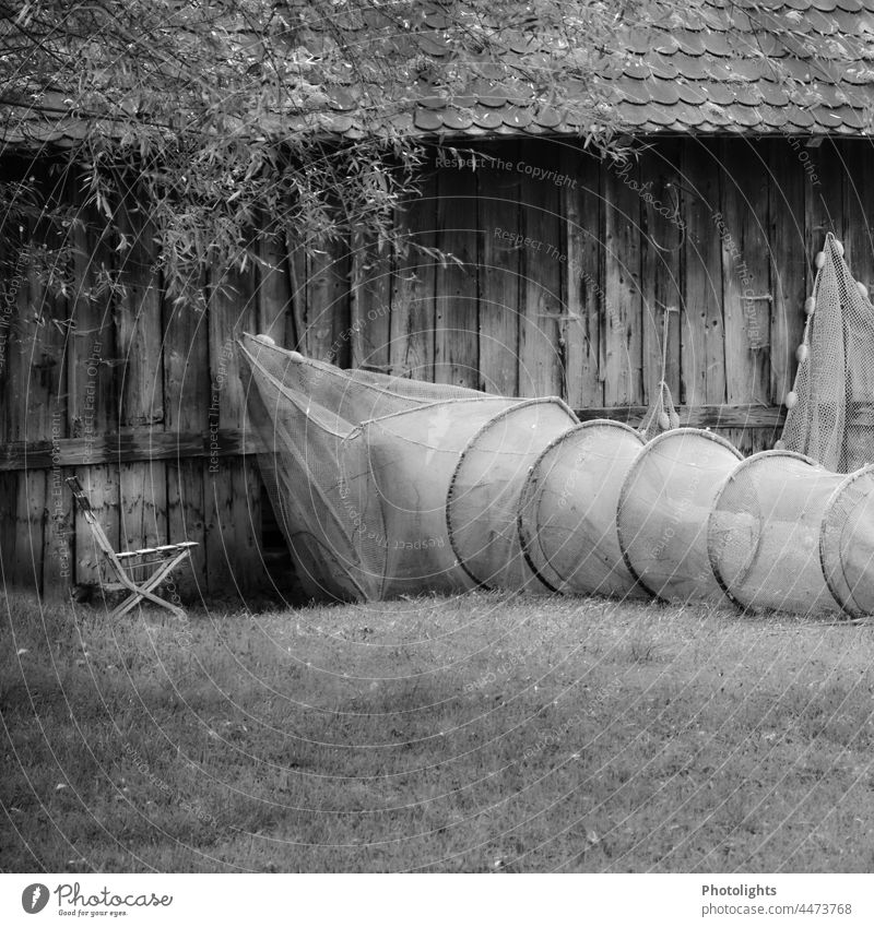 A fishing net hangs on a wooden house wall, next to it lies an eel trap on the meadow with a folding chair next to it. Fish trap Folding chair