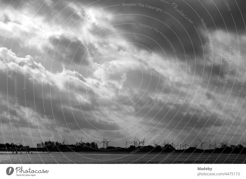 Wind turbines behind the dike at the coast of the North Sea in Bensersiel at the Wadden Sea with wind and clouds in East Frisia in Lower Saxony, photographed in neo-realistic black and white