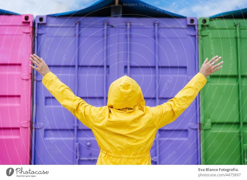 back view of woman with hood and arms raised wearing yellow raincoat over pink and purple background. Colorful Outdoors lifestyle portrait colorful outdoors