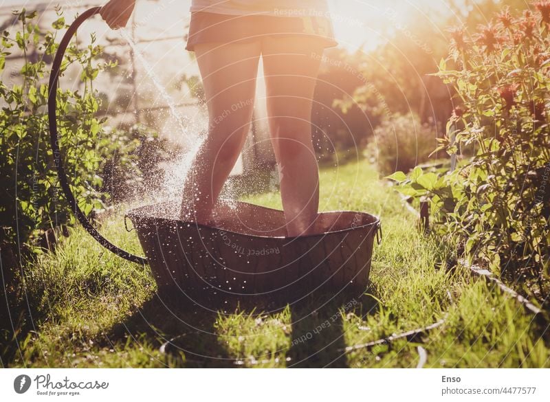Bathing in the summer garden, woman pouring water on her feet from garden hose standing in an iron bath on garden path in sunlight showering bathing legs