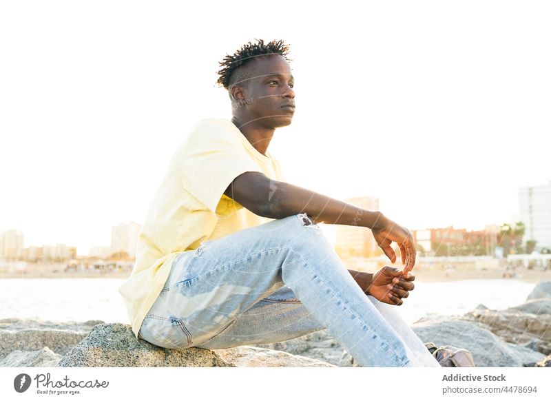 Black man with short hair sitting on shore beach casual serious pensive contemplate calm sea seashore think freedom summer ocean african american blue sky