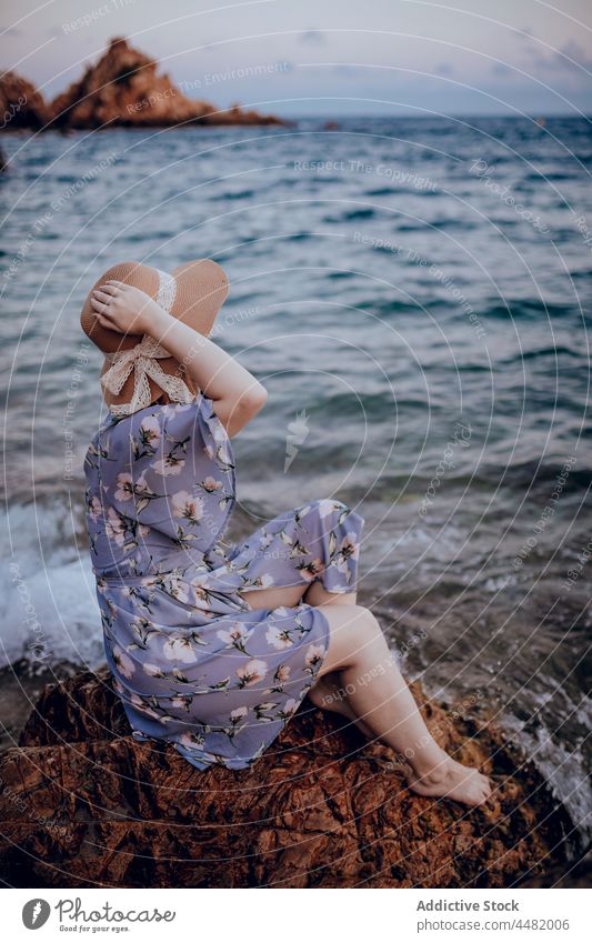 Stylish woman sitting on seashore evening beach coast summer rock dress hat sunset female charming nature water young style stone dreamy calm attractive sundown