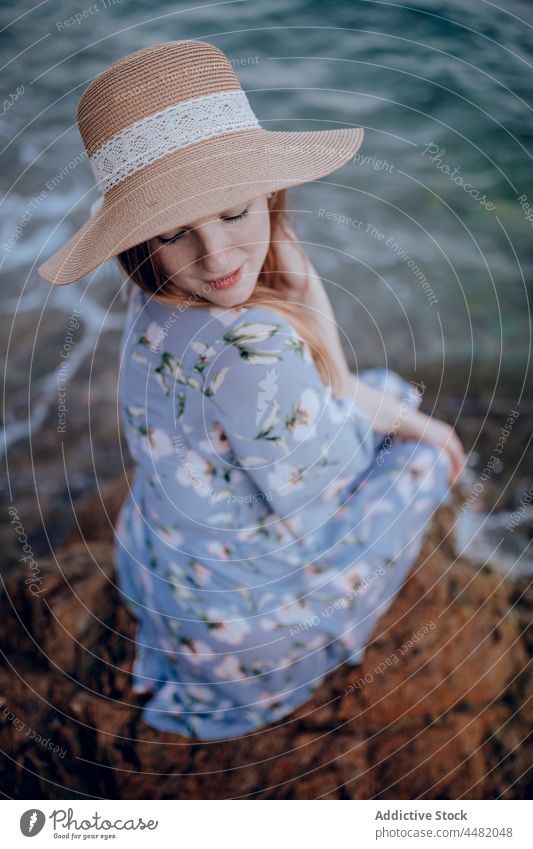 Stylish woman sitting on seashore evening beach coast summer rock dress hat sunset female charming nature water young style stone dreamy calm attractive sundown