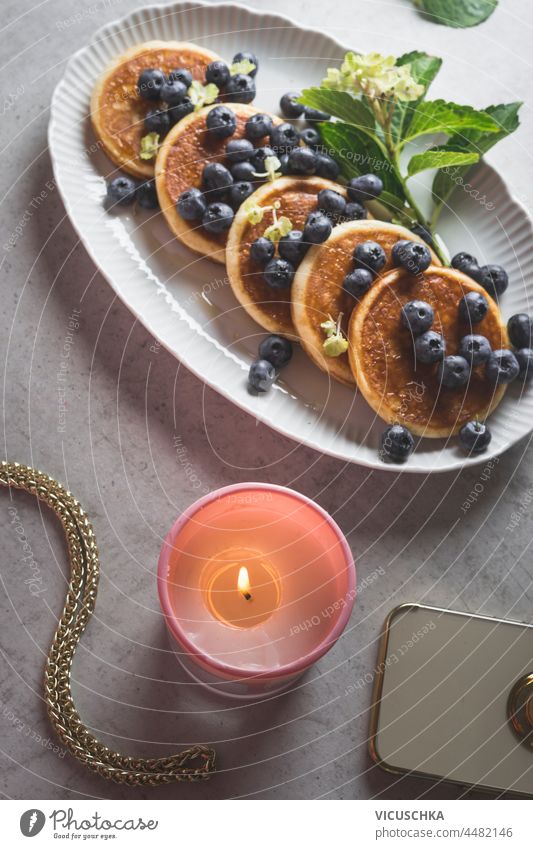 Close up of pancakes with blueberries on white plate, burning candle and smartphone on grey kitchen table. Cozy breakfast with homemade sweet food. Top view.