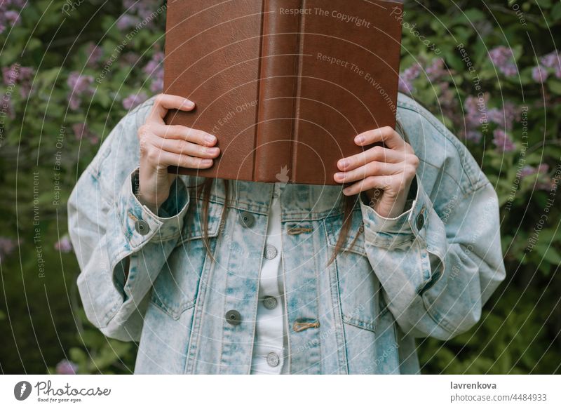 Faceless shot of female holding a book in front of her face outdoors lifestyle young reading portrait education woman intelligent standing green summer hands