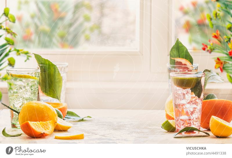 Iced citrus drinks with mint, orange and grapefruit on kitchen table at window with natural light. Homemade healthy lemonade with fruits in summer. Drink background with copy space. Front view.