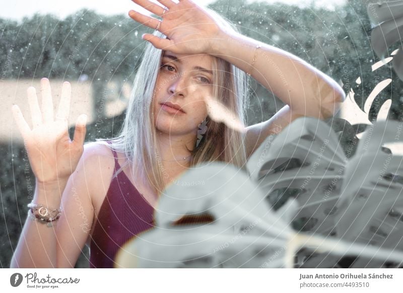 An attractive young woman looking at the camera through glass and surrounded by plants girl pretty lifestyle people garden person white blond hair beauty female