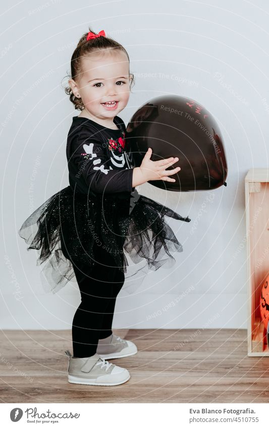 smiling beautiful one year old caucasian girl in halloween costume at home with Halloween decoration, holding balloon. Lifestyle indoors. Halloween party concept.