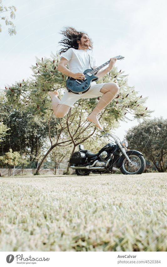 Man jumping while playing a electric guitar with a motorbike as background. Heavy metal lifestyle concept. Relaxed and sunny day at the park. music heavy