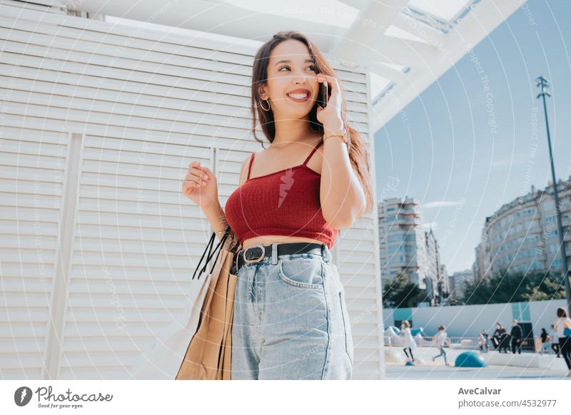 Young arab woman doing a call while holding shopping bags, shopping day, modern outfit styling, smiling during a sunny day mobile phone buying african outside