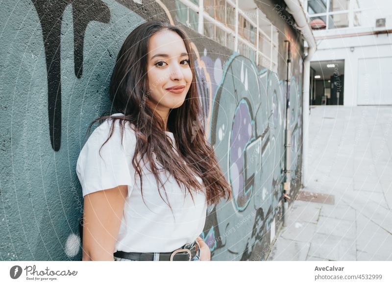 Portrait of young arab woman wearing white shirt and blue jeans smiling to camera on a brick wall with graffiti painting background. Street life style, cool trendy. Social network concept