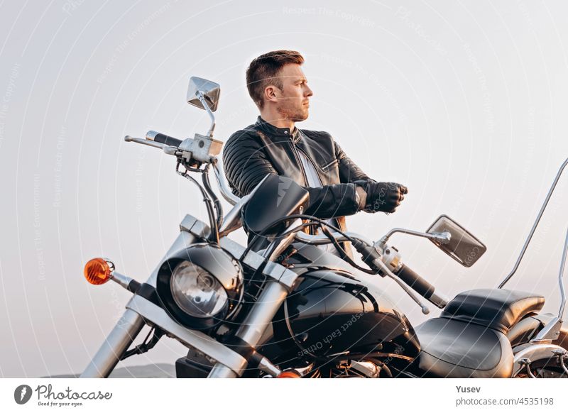 Attractive guy of Caucasian ethnicity in a leather jacket on a motorcycle. Portrait of a handsome young man on a black bike. Summer evening attractive stylish