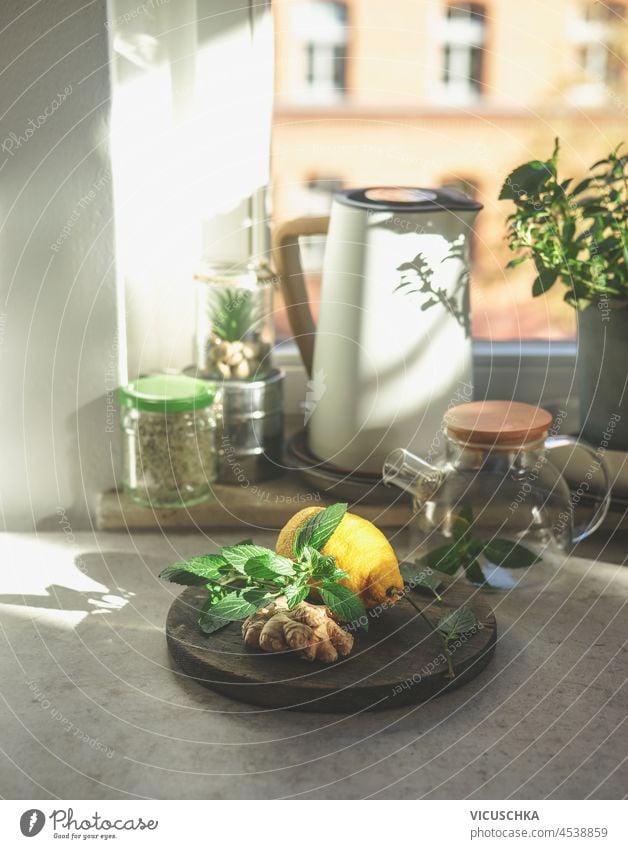 Preparing fresh ginger tea with healthy ingredients at home. Ginger, mint leaves and lemon on kitchen table with glass tea pot, electric kettle and jars at window background.  Front view.