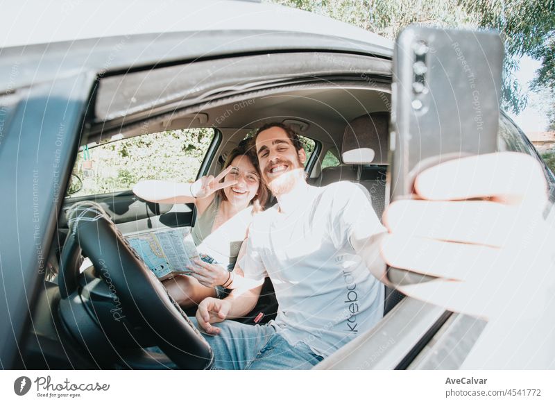 Lovely young couple traveling doing a road trip in car. Road travel, frontal portrait smiling. They are taking a break from driving and taking a selfie with their phone. Hipster modern couple.