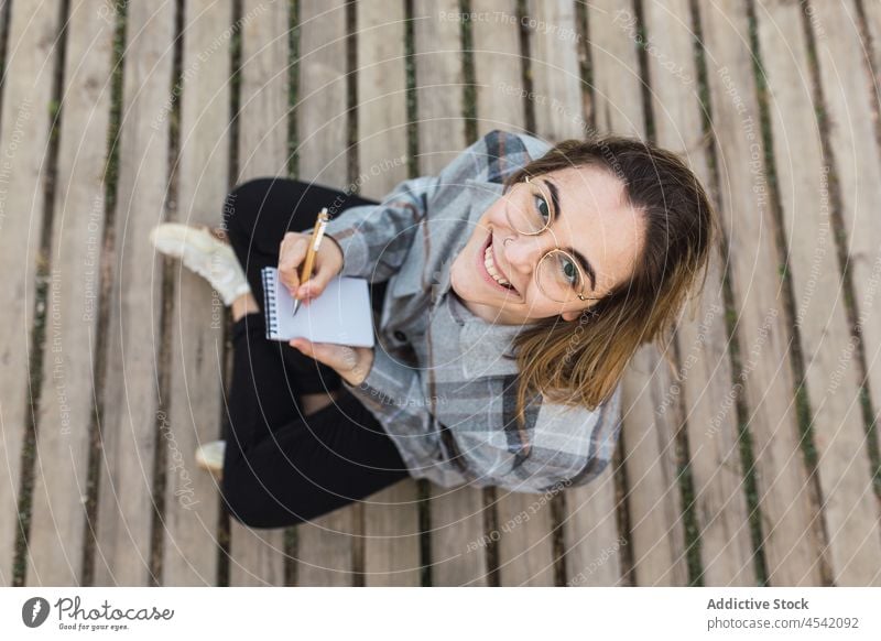 Dreamy woman sitting on boardwalk and writing in notebook in nature take note path smile write dreamy inspiration writer sea female young casual eyeglasses